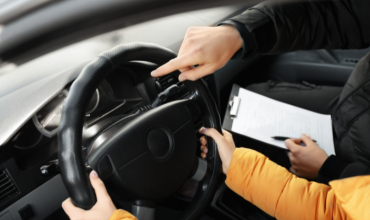 Twee mensen zitten in de auto. De rij-instructuur, die een klembord vasthoudt, wijst naar het stuur van de bestuurder.