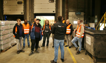 Een groep mensen in veiligheidshesjes luistert naar een man die een trui met 'Vos Logistics' erop draagt.